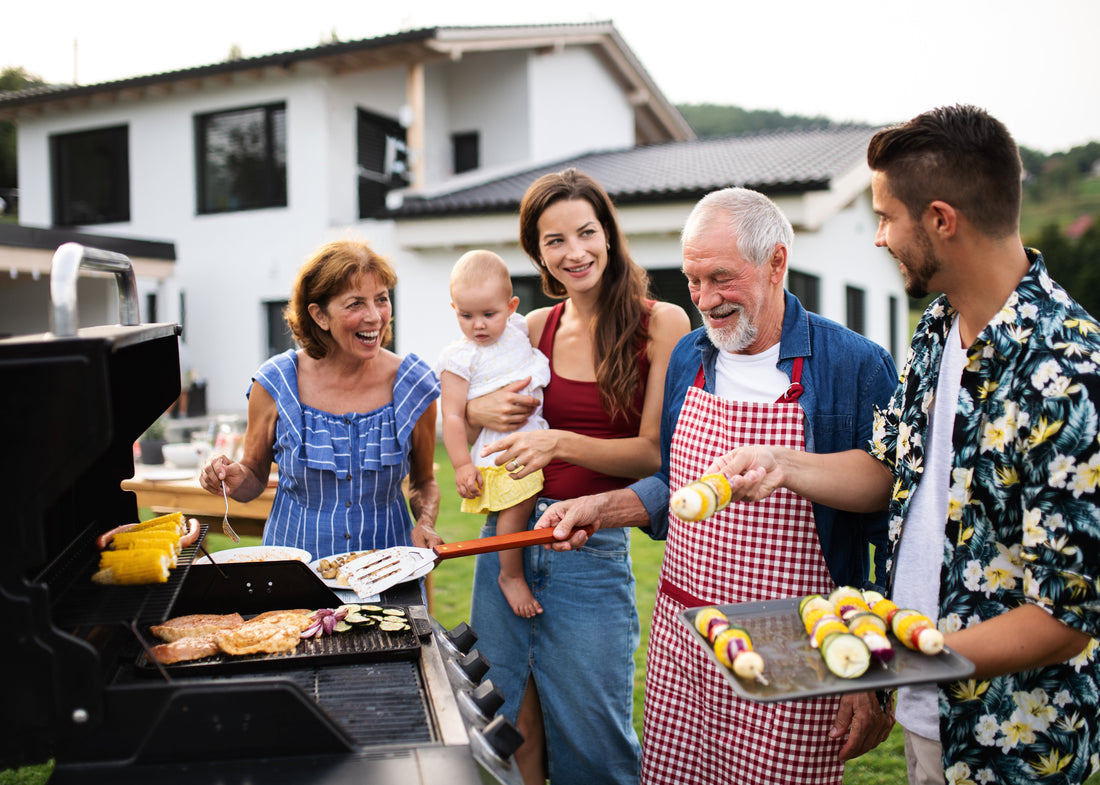 Family barbecuing in a backyard. An older man in a red-checkered apron grills food. Others, including a baby, smile and chat. Modern house background.