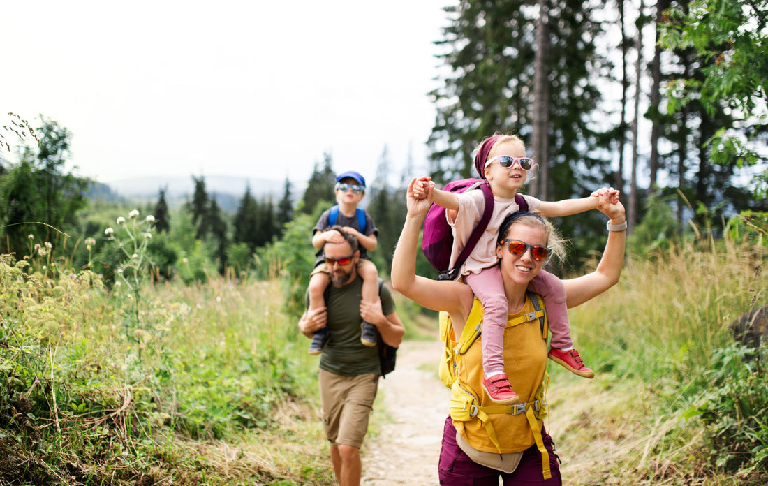 Family hiking in a forest, children on parents' shoulders. Bright clothing and sunglasses, lush greenery, creating a fun, adventurous mood.