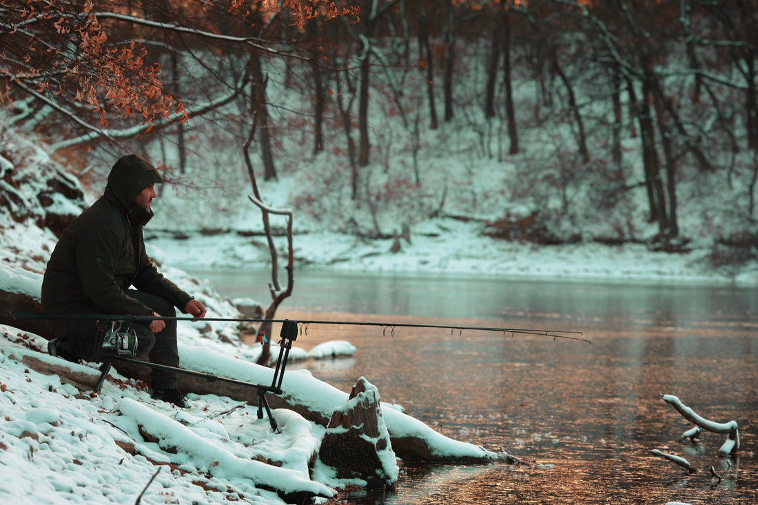 A man fishing on a river bank in the snow.