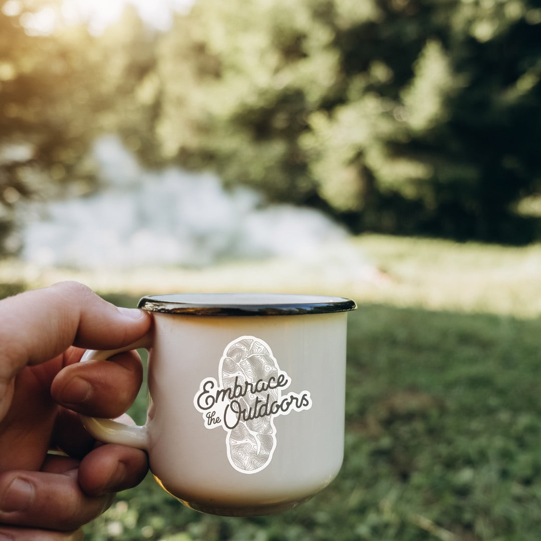 Person holding a mug with 'Embrace the Outdoors' text against a natural background